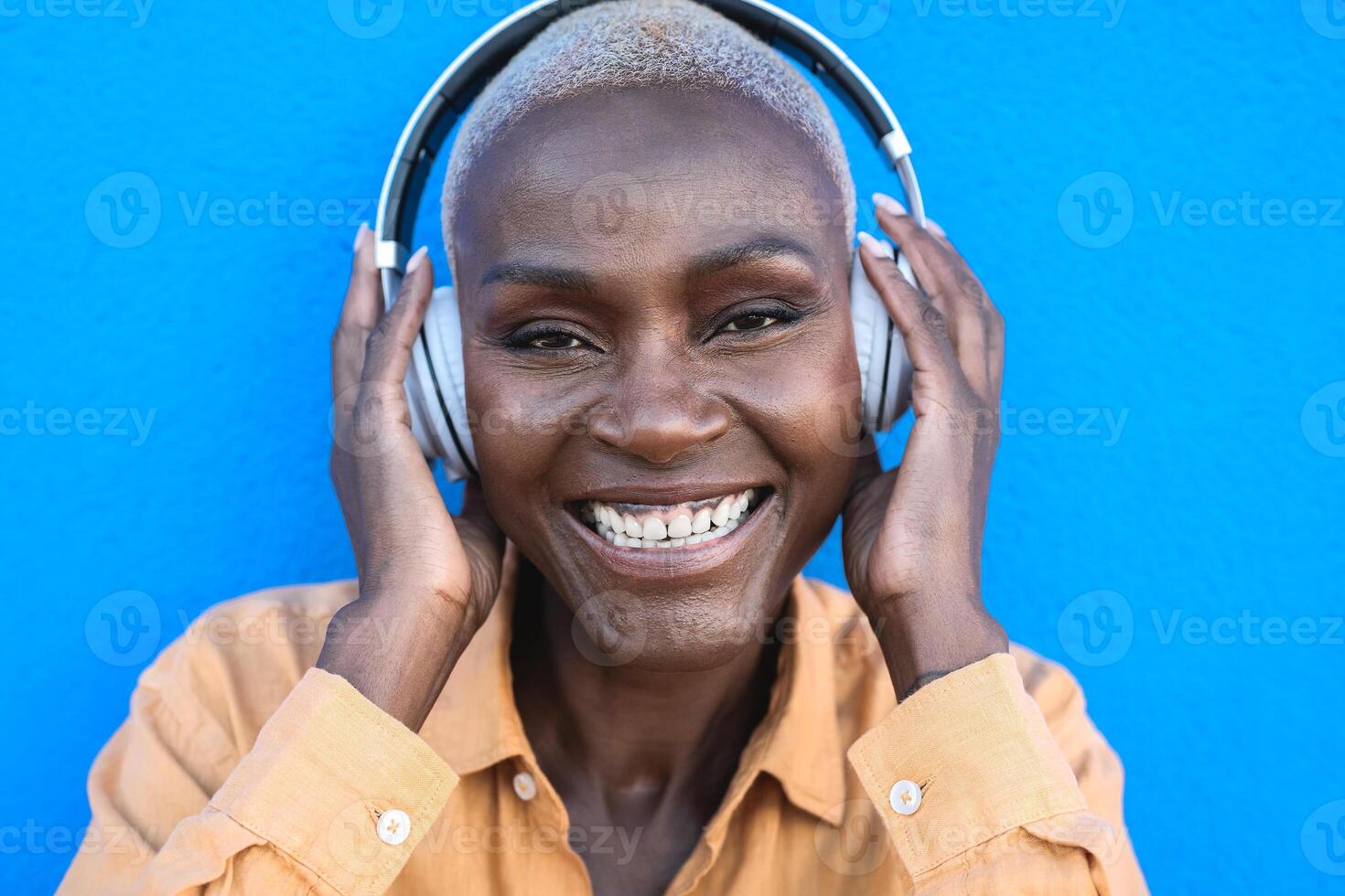 contento anziano africano donna avendo divertimento ascoltando musica con senza fili cuffie contro blu sfondo foto