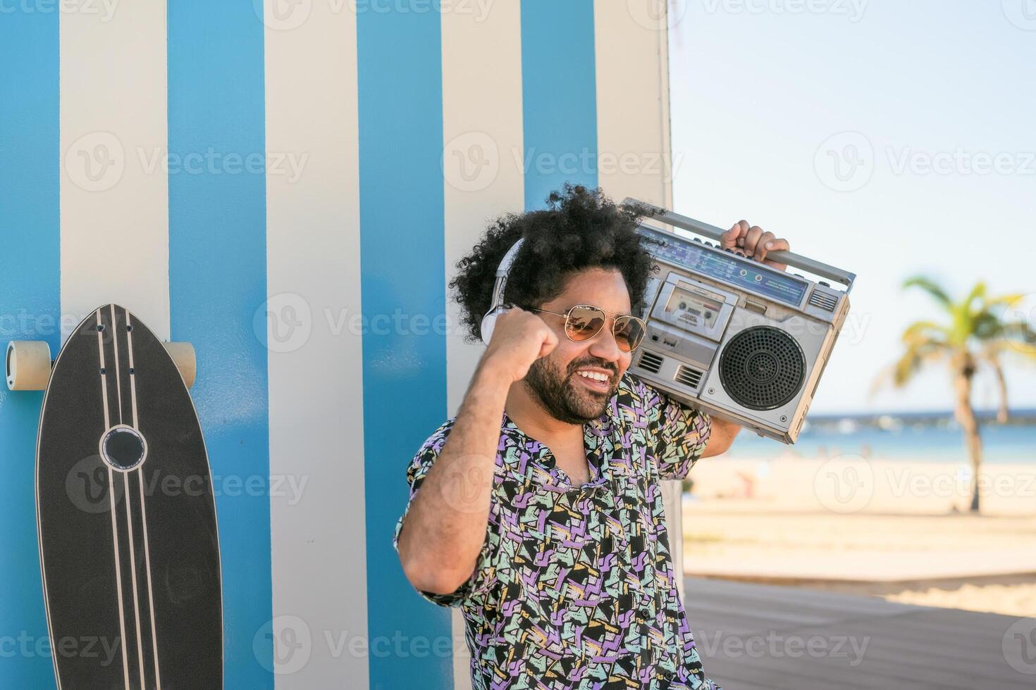 contento afro uomo avendo divertimento ascoltando per musica con senza fili cuffie e Vintage ▾ Boombox su il spiaggia durante estate tempo foto