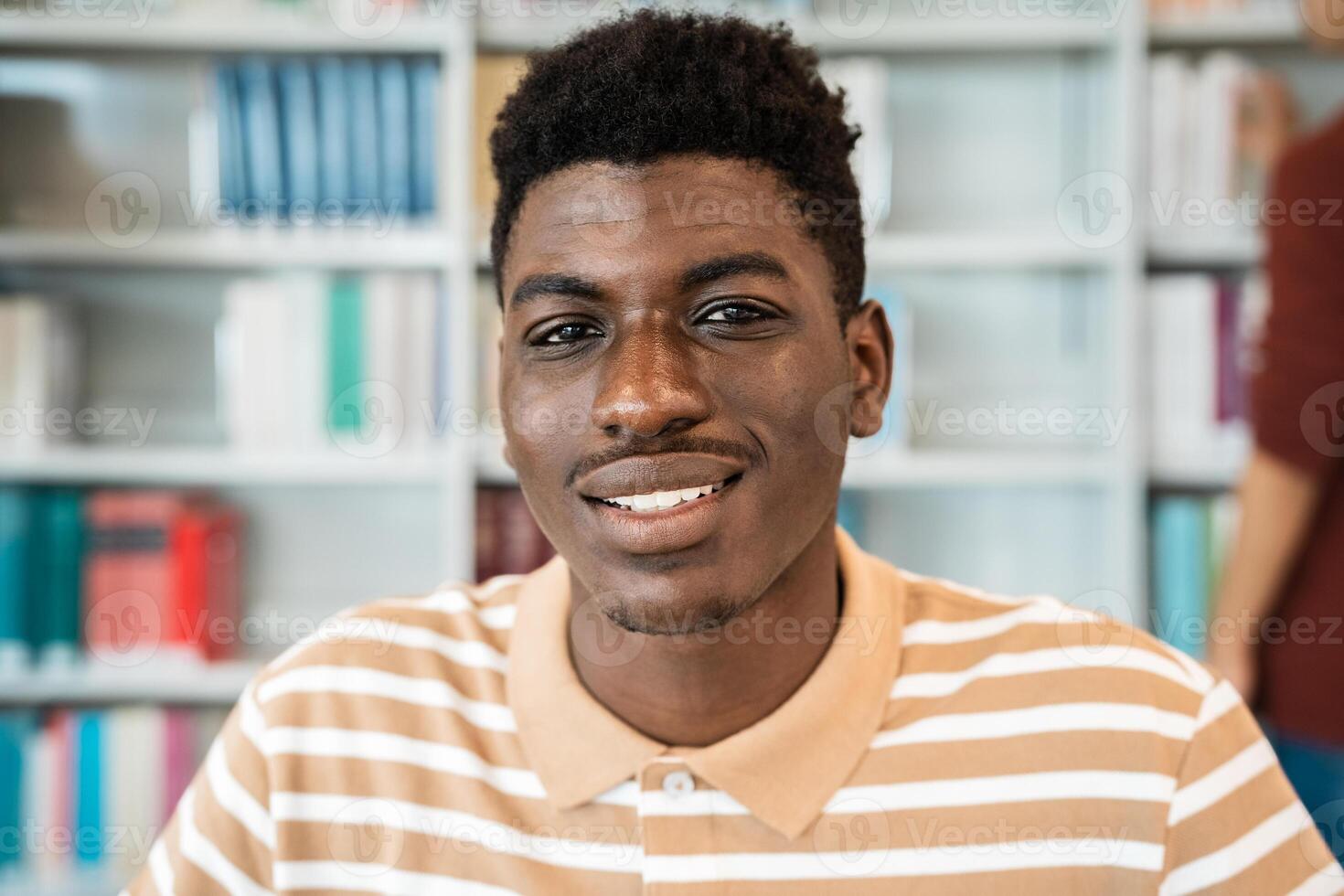 contento giovane africano uomo sorridente in il telecamera mentre in piedi dentro Università biblioteca foto