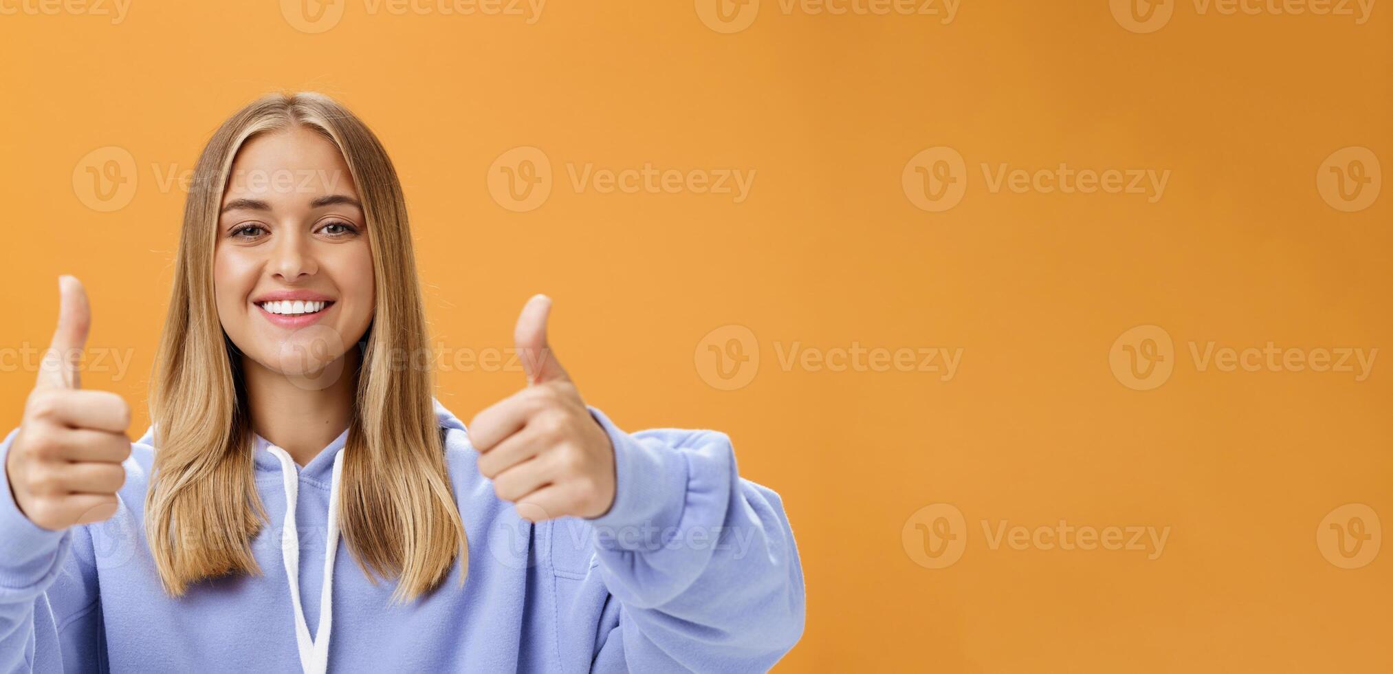 ritratto di contento ottimista donna mostrando pollici su gesto e sorridente supporto con allegro Guarda approvazione e simpatia grande Prodotto dando positivo opinione e raccomandazione al di sopra di arancia parete foto