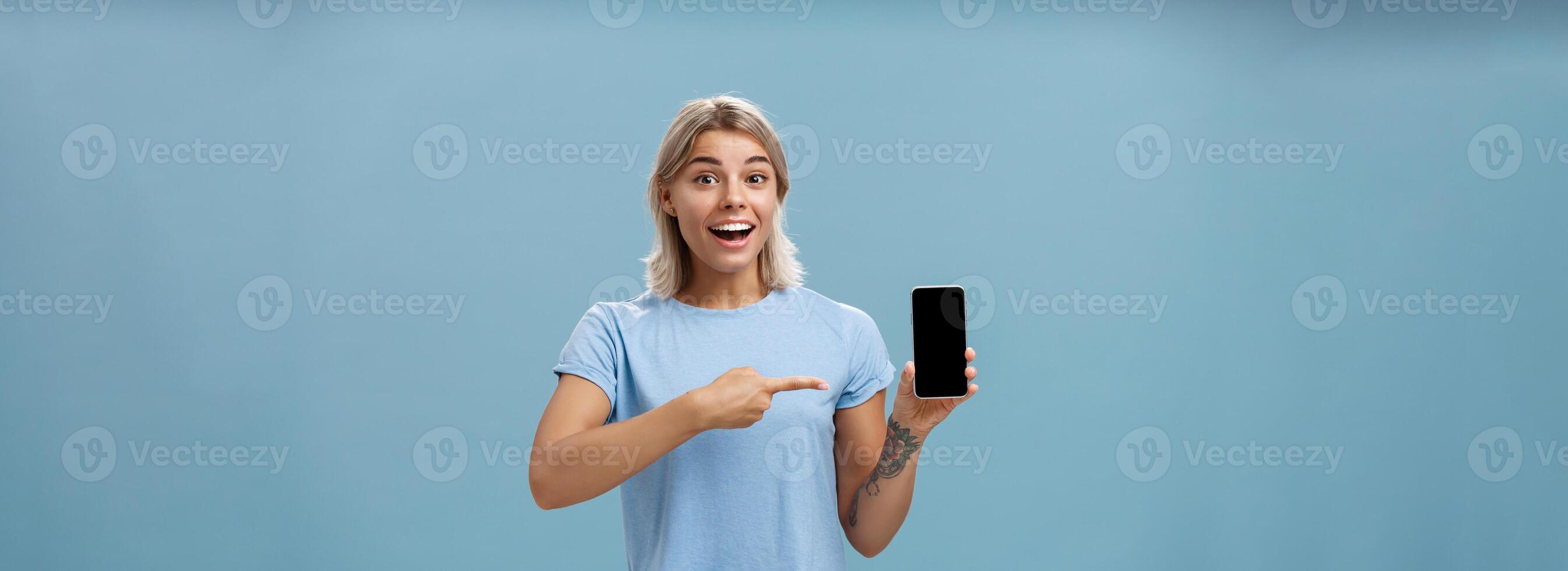 dalla vita in su tiro di entusiasta e impressionato bella vista femmina alunno nel casuale maglietta sorridente con gioia puntamento a smartphone schermo mostrando eccezionale posto attraverso Internet per amico al di sopra di blu sfondo foto