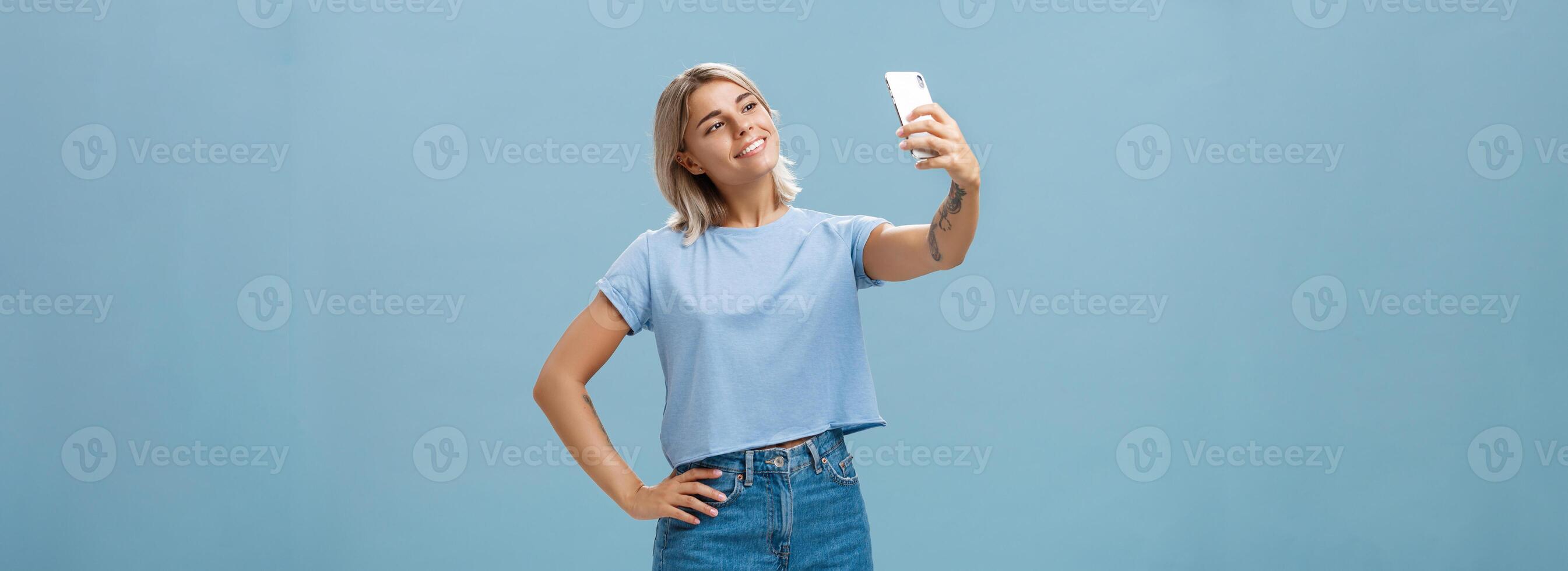 elegante carino femmina Palestra manager assunzione autoscatto con nuovo smartphone in posa con ampio lieta Sorridi e Tenere mano su anca fabbricazione inviare per seguaci in piedi spensierato e rilassato al di sopra di blu parete foto