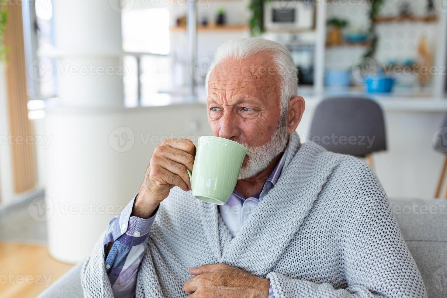 anziano uomo sofferenza a partire dal influenza potabile tè mentre seduta avvolto nel un' coperta su il divano a casa. malato più vecchio uomo con mal di testa seduta sotto il coperta nel il vivente camera. uomo con un' freddo foto