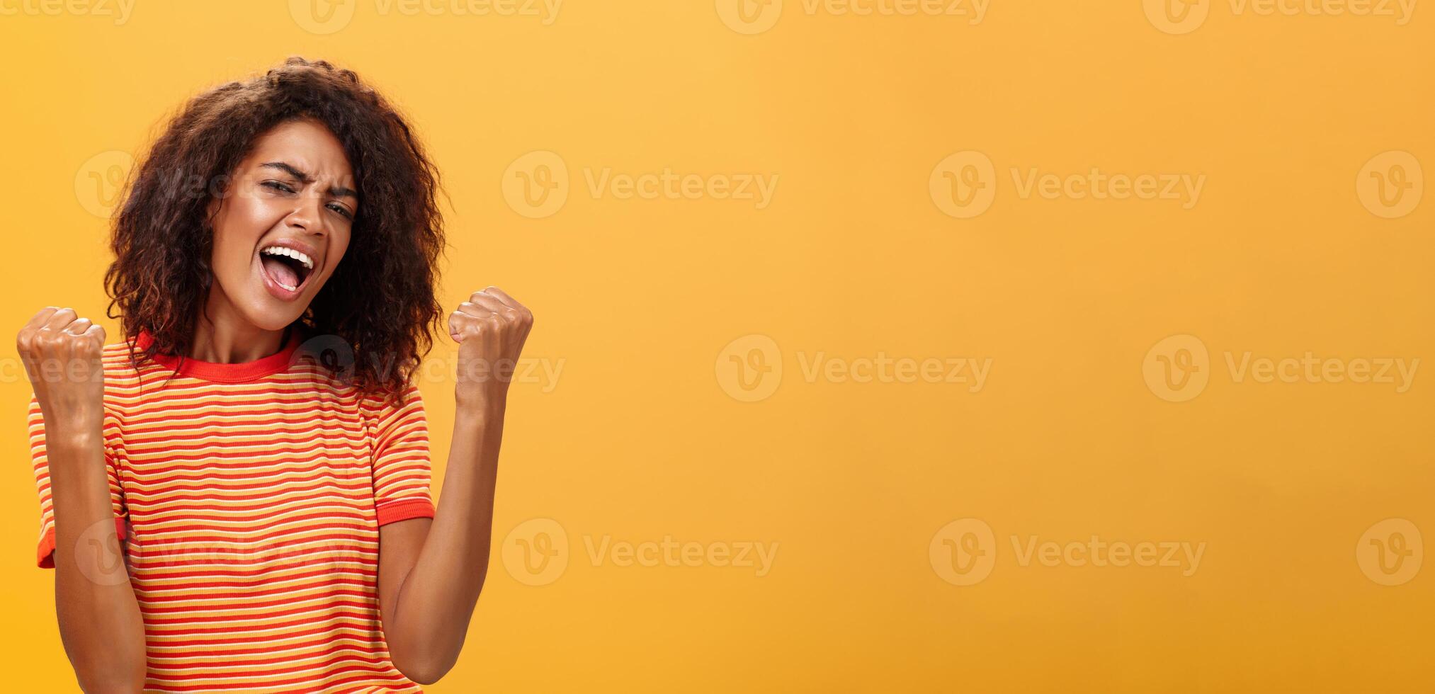 allegro Contentissimo e entusiasta africano americano donna con afro acconciatura stringendo sollevato pugni urla sì a partire dal trionfo e gioia di successo in piedi soddisfatto di vittoria al di sopra di arancia parete foto