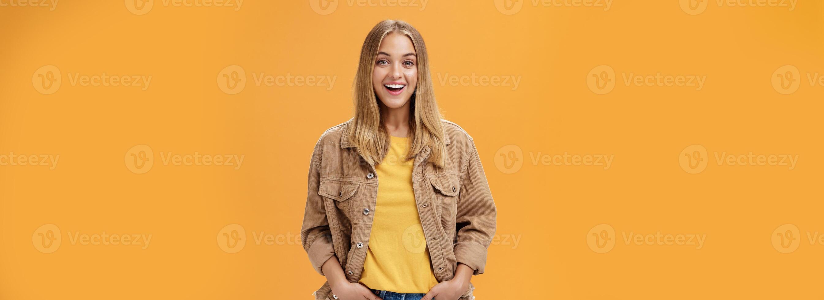 carismatico abbronzato donna nel velluto a coste giacca e giallo maglietta pronto per freddo autunno camminare con amici sorridente con gioia guardando intrattenuto a telecamera Tenere mano nel tasche casualmente al di sopra di arancia parete foto