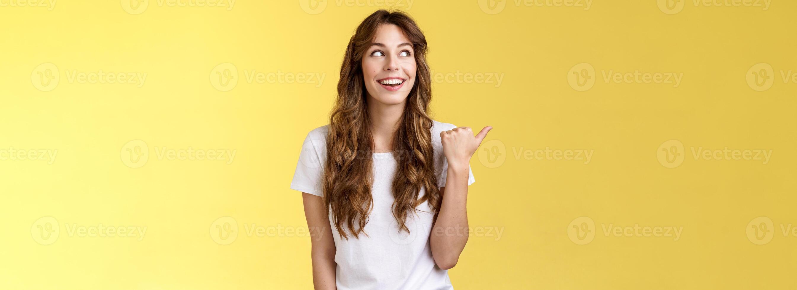 qualcosa interessante giusto là. curioso vivace entusiasta affascinante donna guardando puntamento pollice sinistra sorridente a grandi linee discutere freddo evento ridendo spensierato In piedi giallo sfondo foto