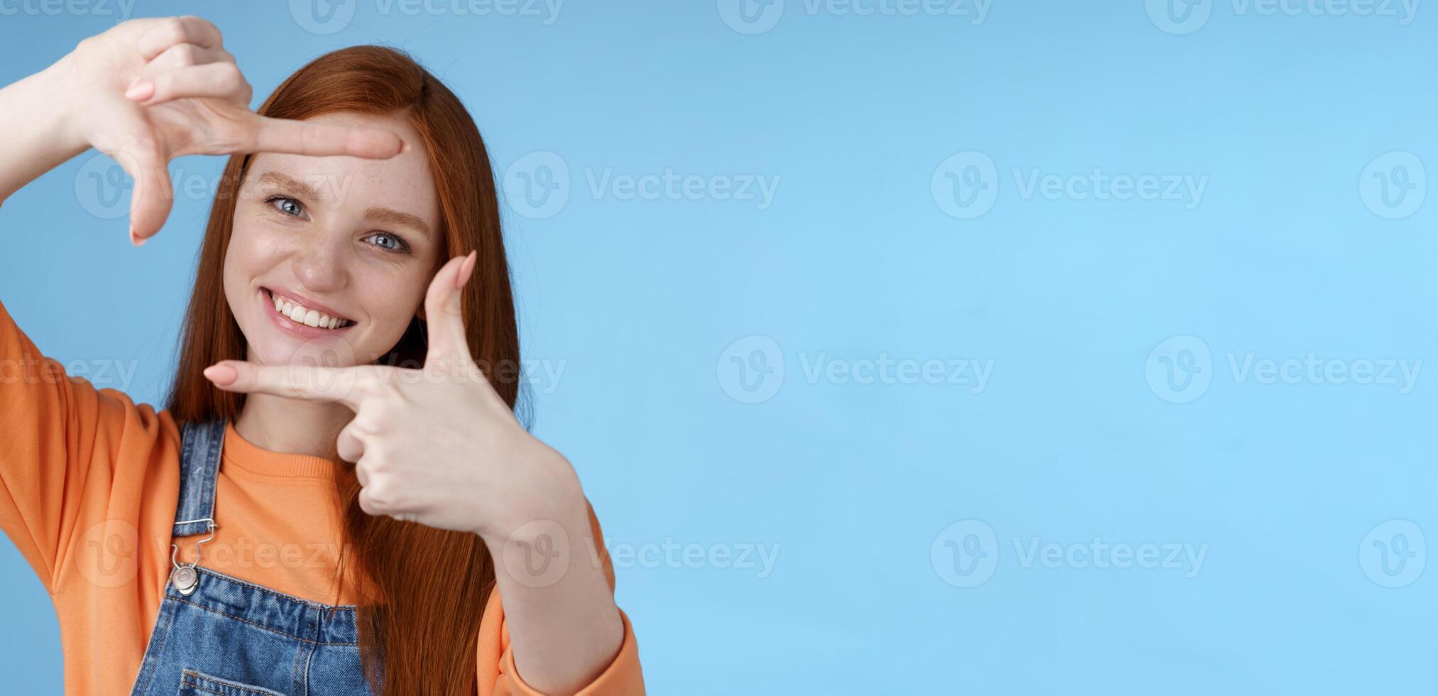 gioioso attraente sincero testa Rossa giovane ragazza ricerca ispirazione trova Perfetto angolo prendere bene tiro rendere mano montatura Guarda attraverso Contentissimo divertito sorridente a grandi linee bianca denti, blu sfondo foto