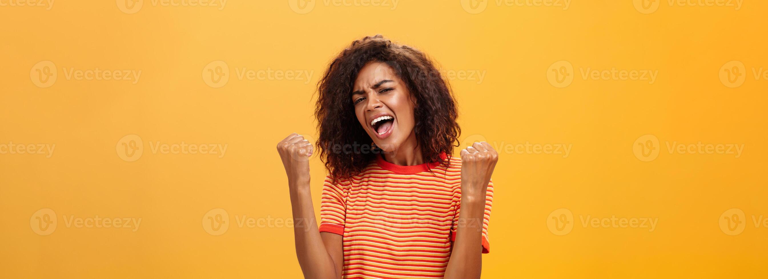 allegro Contentissimo e entusiasta africano americano donna con afro acconciatura stringendo sollevato pugni urla sì a partire dal trionfo e gioia di successo in piedi soddisfatto di vittoria al di sopra di arancia parete foto