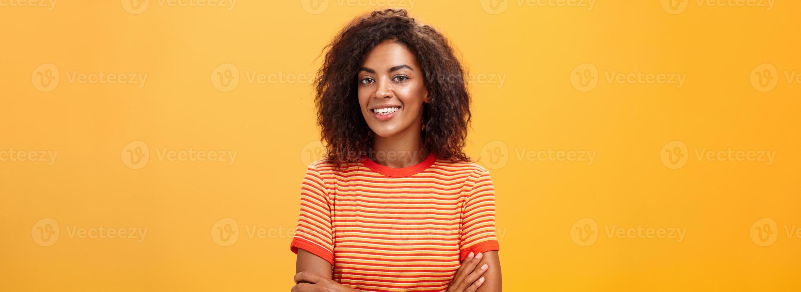 dalla vita in su tiro di creativo ambizioso bella vista femmina collaboratore nel di moda a strisce maglietta Tenere mani attraversato su il petto sorridente a grandi linee a telecamera amichevole e sicuro di sé al di sopra di arancia sfondo foto