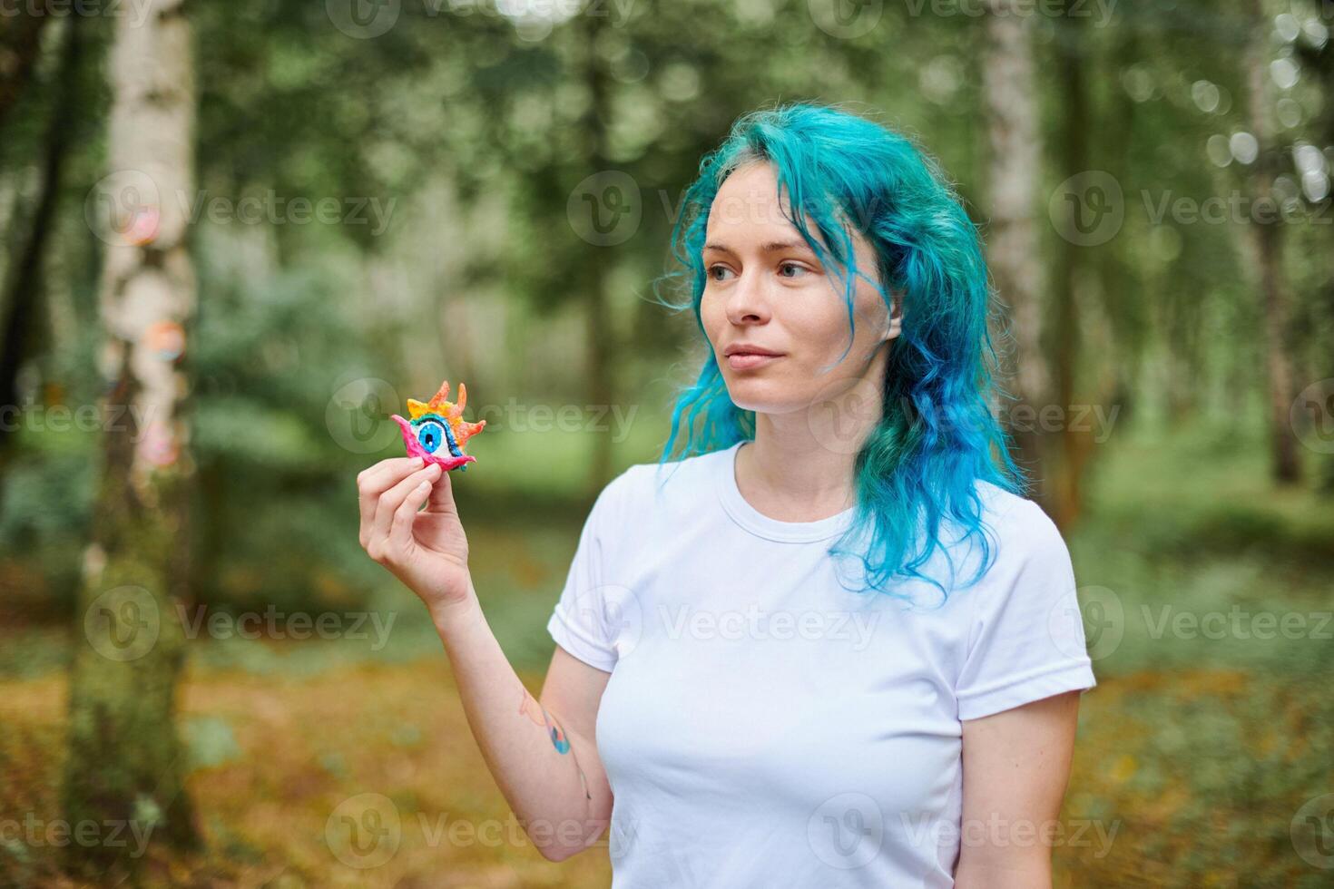 giovane donna con turchese tinti capelli nel bianca t camicia detiene fatto a mano colorato occhio amuleto nel parco foto