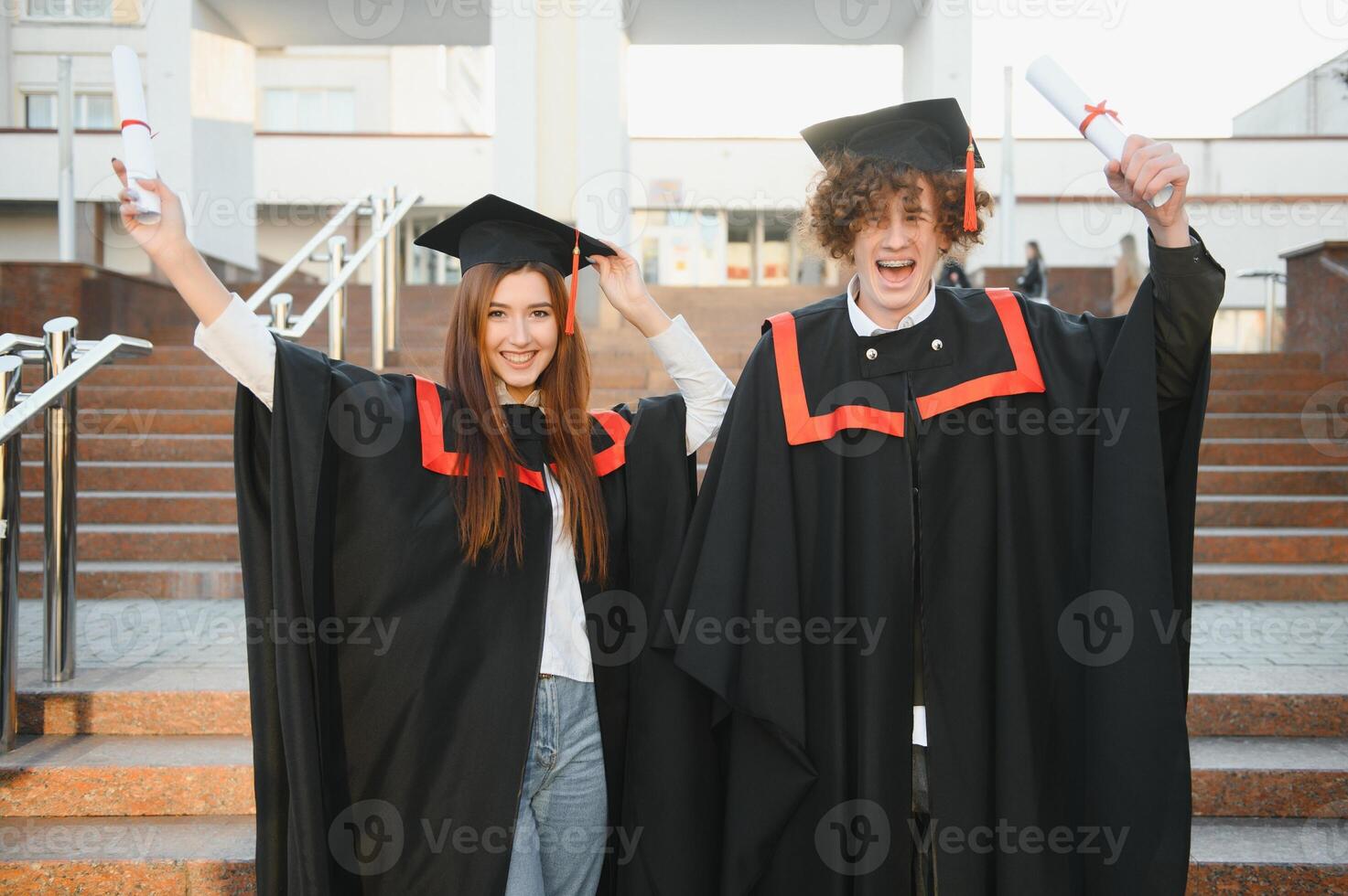 ritratto di contento laureati. Due amici nel la laurea caps e abiti in piedi al di fuori Università edificio con altro studenti nel sfondo, Tenere diploma pergamene, e sorridente foto