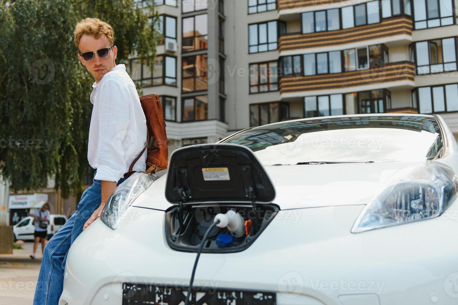 uomo ricarica il suo lusso elettrico auto a all'aperto stazione nel davanti di moderno nuovo città edifici foto