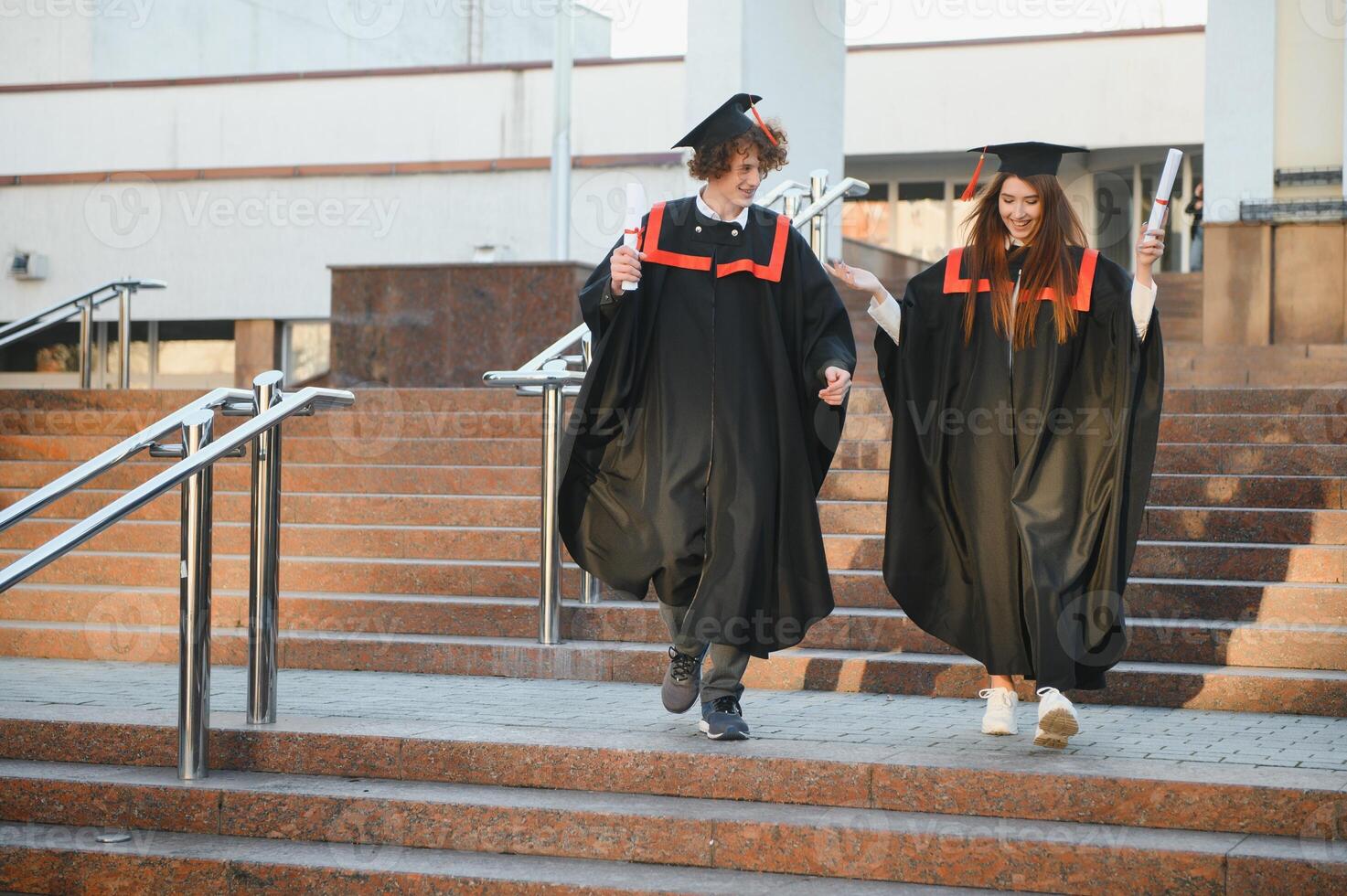 ritratto di contento laureati. Due amici nel la laurea caps e abiti in piedi al di fuori Università edificio con altro studenti nel sfondo, Tenere diploma pergamene, e sorridente foto