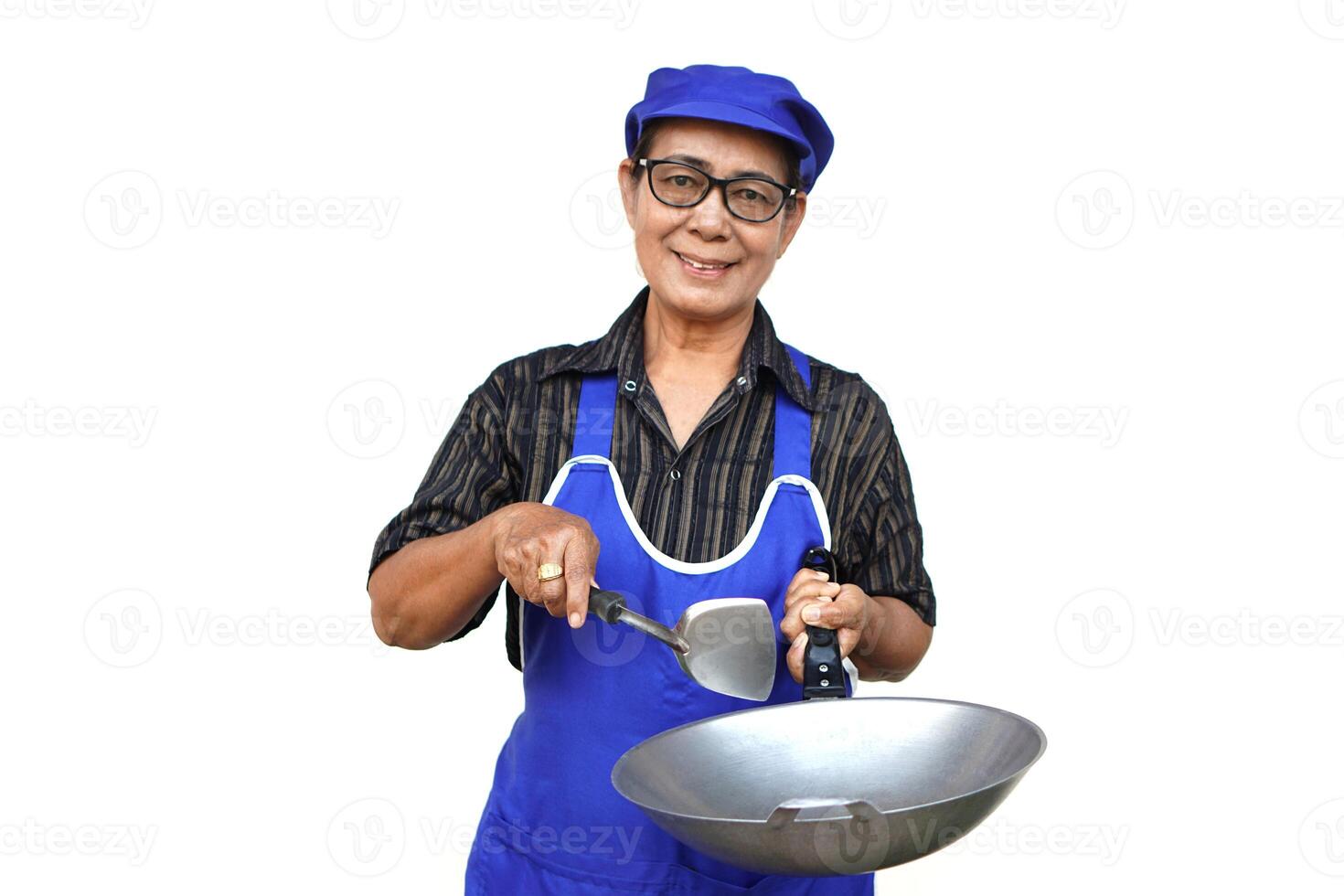 asiatico anziano donna, indossa blu grembiule e berretto, detiene frittura padella e spatola o mestolo. Guarda a telecamera, sorrisi, si sente fiducioso isolato su bianca sfondo. concetto, amore cucinando. cucina stile di vita. foto