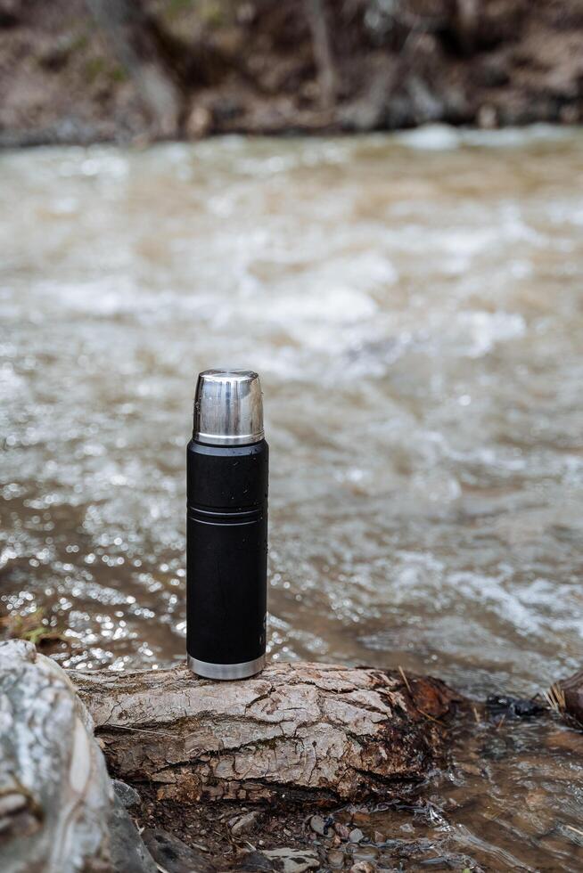 un' thermos sta su il sfondo di acqua, un' nero vuoto bottiglia, un' termo boccale per caldo bevande. foto