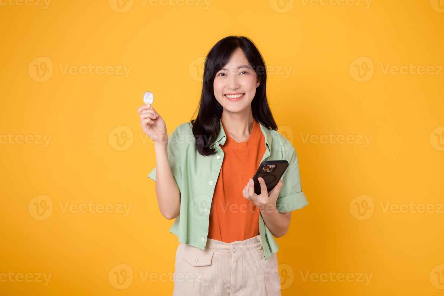 futuro di finanza con un' allegro giovane asiatico donna anni '30, indossare arancia camicia e verde Maglione, visualizzazione crypto moneta moneta mentre Tenere smartphone su giallo sfondo. futuro finanza concetto. foto