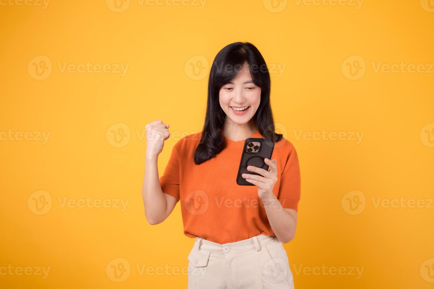 allegro asiatico donna nel anni '30, indossare arancia camicia, utilizzando smartphone con cazzotto su mano cartello su vivace giallo sfondo. celebrare nuovo App concetto. foto