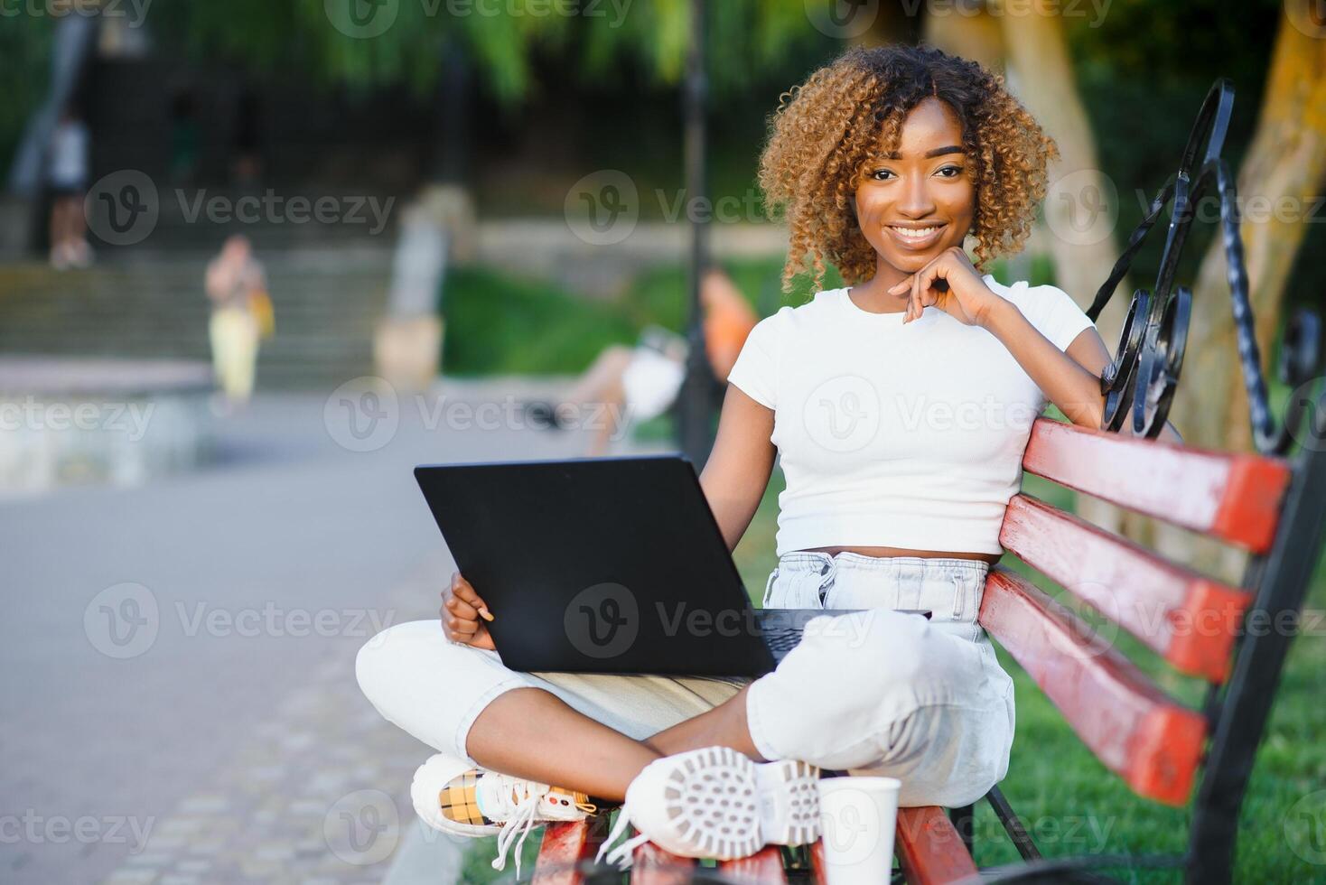 bellissimo giovane misto gara donna Lavorando con il computer portatile e utilizzando Telefono. seduta su il panchina nel parco, all'aperto ritratto , indossa casuale Abiti e bicchieri foto