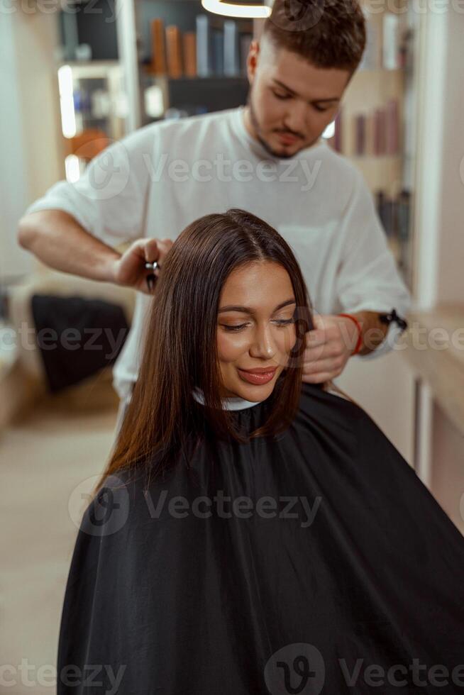 un' contento giovane donna si siede nel un' sedia coperto con un' capo mentre un' capelli maestro tagli sua capelli foto