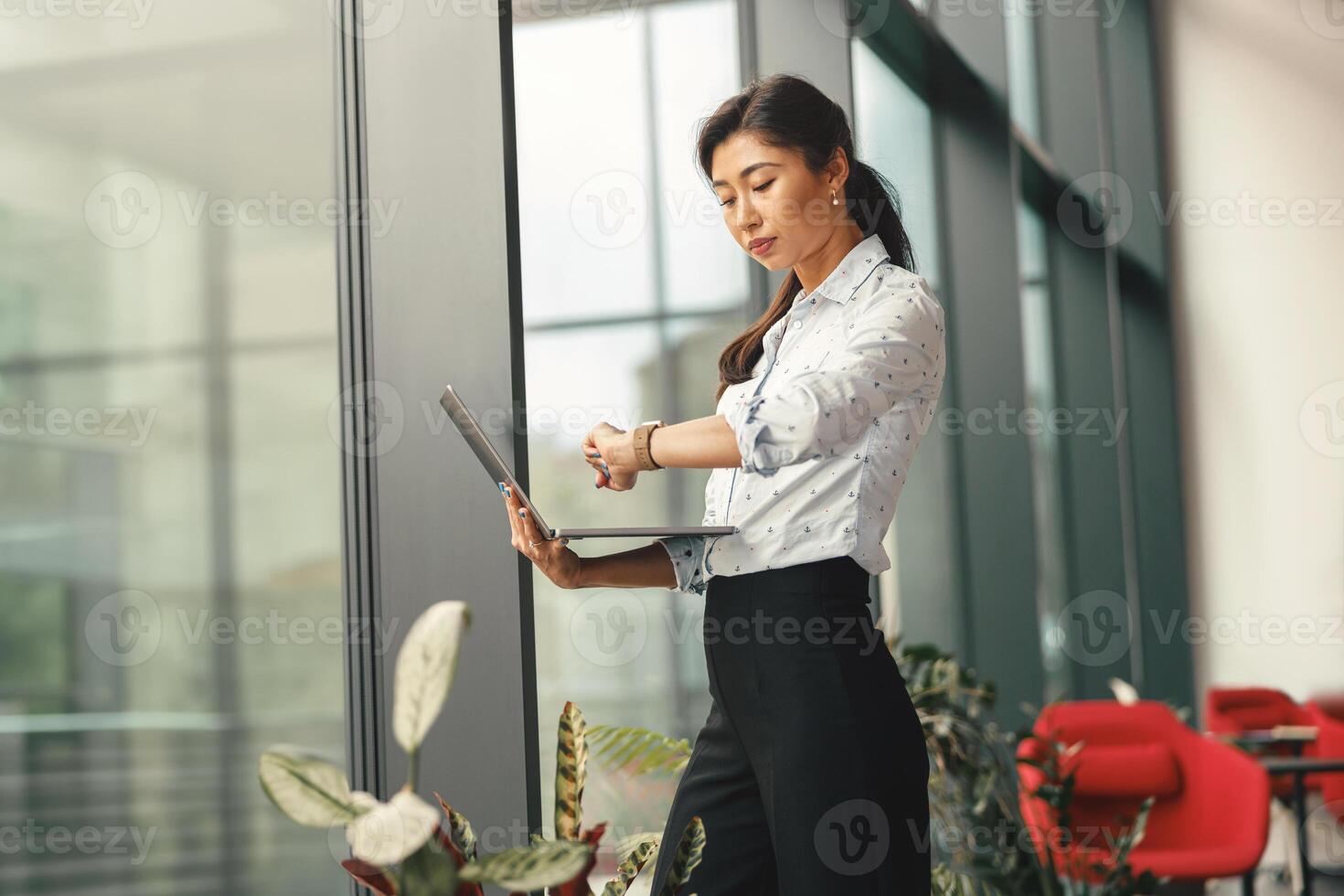 focalizzata donna d'affari guardare su sua polso orologio mentre in piedi con il computer portatile vicino ufficio finestra foto