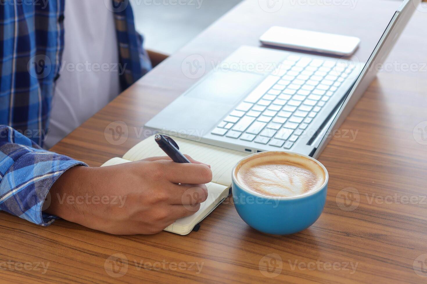 vicino su di maschio mano scrittura su Nota libro con penna, mostrando vuoto schermo di il computer portatile e smartphone. Lavorando nel bar concetto con un' tazza di caffè foto