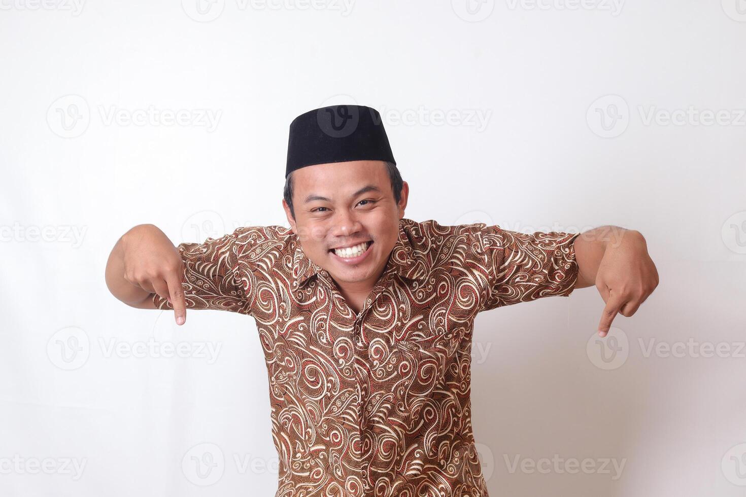 ritratto di eccitato asiatico uomo indossare batik camicia e songkok sorridente e guardare a il telecamera puntamento con Due mani e dita per il giù lato. isolato Immagine su grigio sfondo foto