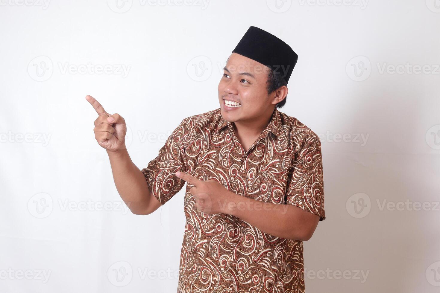 ritratto di eccitato asiatico uomo indossare batik camicia e songkok sorridente e guardare a il telecamera puntamento con Due mani e dita per il lato. isolato Immagine su grigio sfondo foto