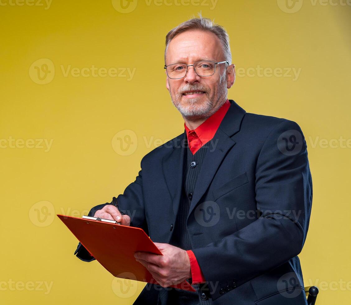 anziano uomo d'affari seduta con cartella al di sopra di giallo sfondo nel completo da uomo guardare a telecamera nel rosso camicia . attività commerciale concetto foto