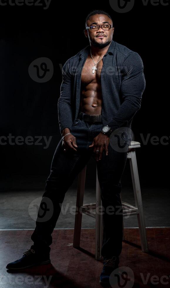 pieno lunghezza Visualizza di il africano americano bodybuilder in piedi con fiducia su nero studio sfondo. uomo dimostrando il suo stampa e muscoli foto