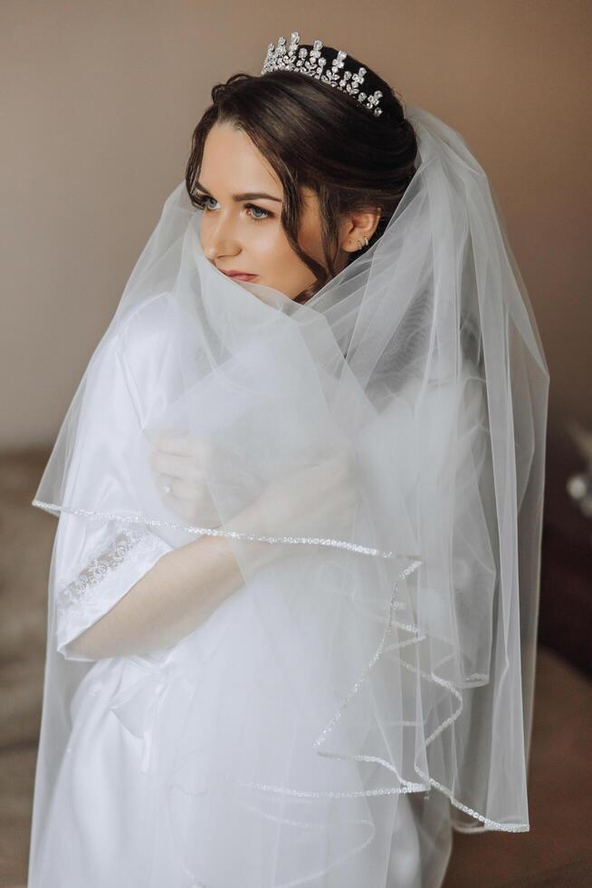 ritratto di un incredibilmente bellissimo ragazza sposa nel un' bianca accappatoio nel il Camera da letto, il sposa pose Tenere sua velo nel sua mani e coperture se stessa con esso. foto