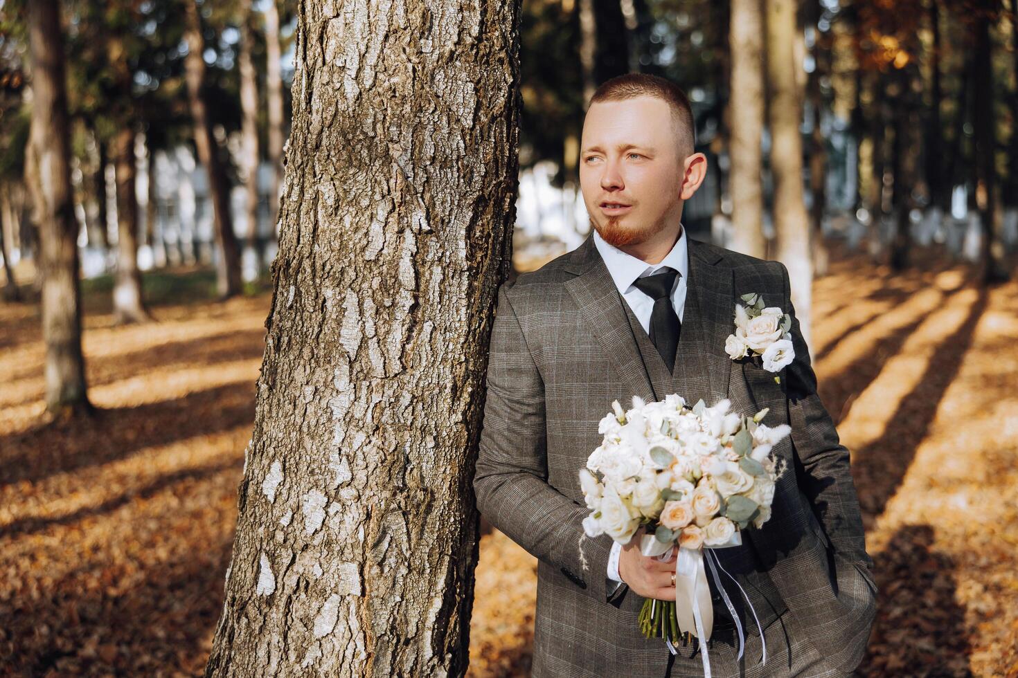 ritratto di un' bello giovane sposo su un autunno giorno all'aperto nel un elegante completo da uomo e Tenere un' nozze mazzo di fiori. all'aperto foto. bello uomo nel un' attività commerciale completo da uomo. foto