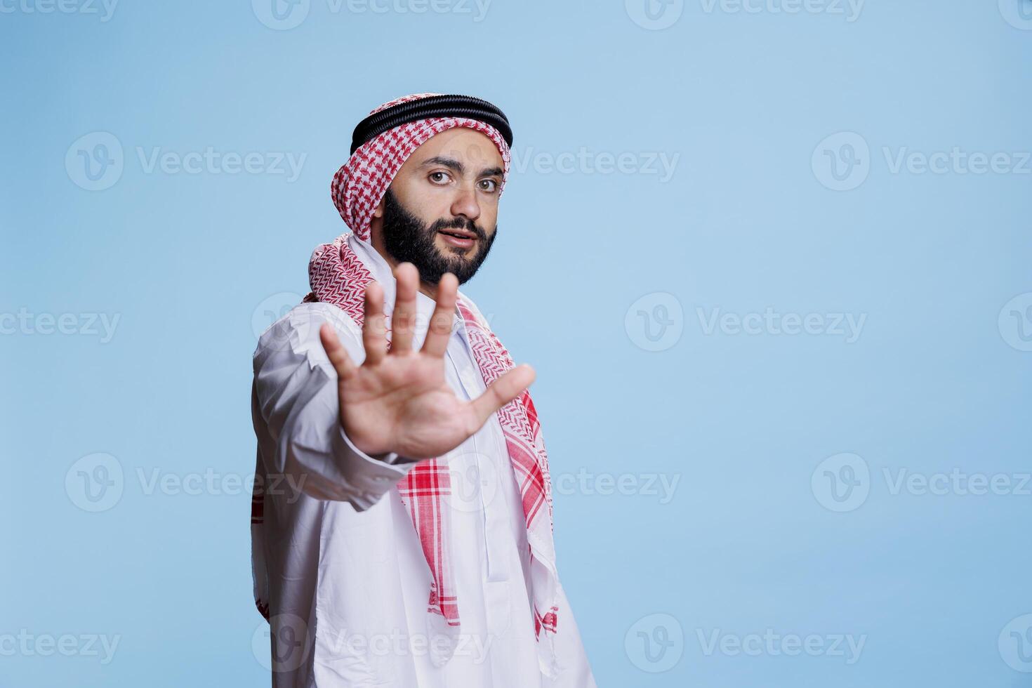 musulmano uomo indossare tradizionale Abiti mostrando fermare gesto con mano e guardare a telecamera con grave espressione. fiducioso arabo in posa con divieto cartello studio ritratto foto