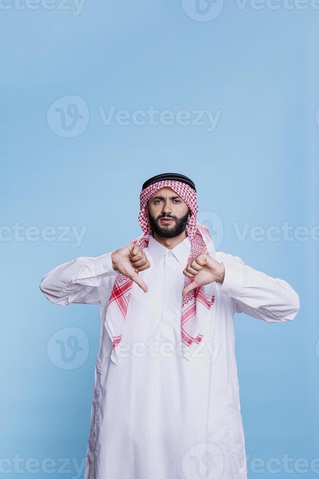 malcontento arabo indossare tradizionale Abiti in posa con pollici giù e guardare a telecamera con grave espressione. musulmano uomo vestito nel thobe e ghutra e mostrando disapprovazione gesto foto