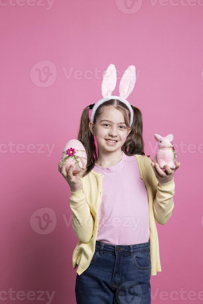 gioioso bambino piccolo presentazione un' rosa uovo e un' ripieno coniglio nel studio, indossare trecce e carino coniglietto orecchie su telecamera. sorridente giovane ragazzo mostrando sua Pasqua decorazioni e colorato giocattoli. foto