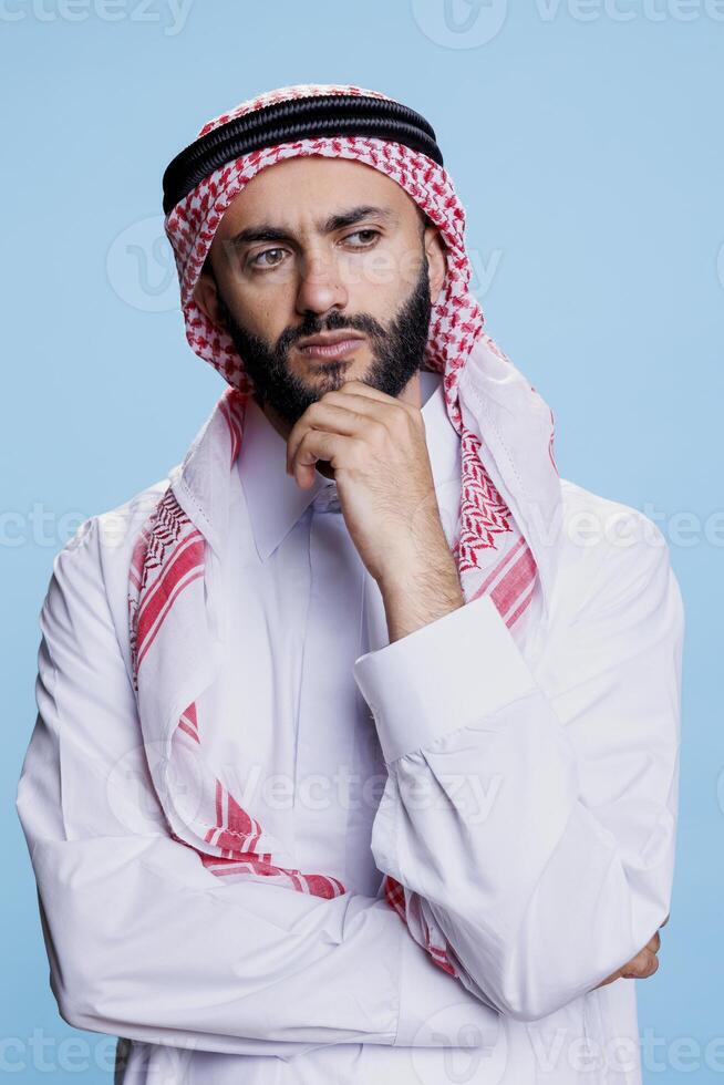 musulmano uomo con nel tradizionale abbigliamento contemplando con pensieroso Guarda. interrogato arabo persona indossare thobe e ghutra foulard in posa nel pensiero posa con mano supporto mento foto