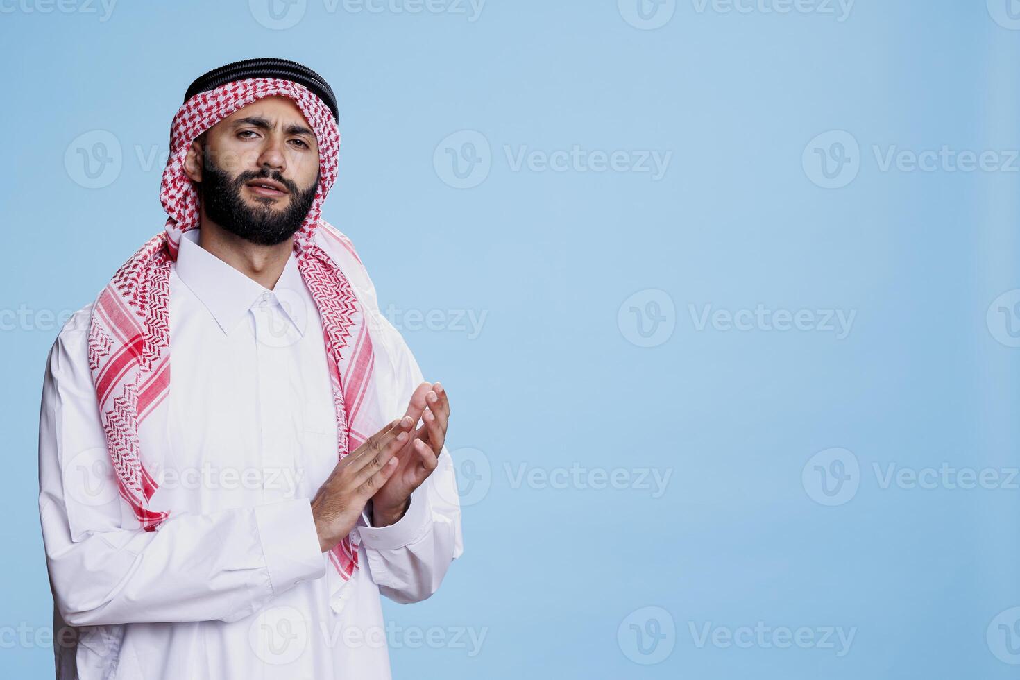 musulmano uomo vestito nel tradizionale accappatoio in mostra orgoglioso espressione mentre Applaudire con mani nel studio. arabo indossare bianca thobe e scacchi ghutra battimani braccia e guardare a telecamera foto