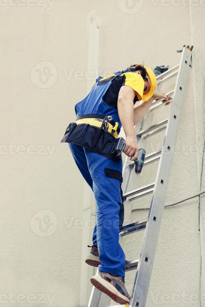 esperto tecnico addetto alla manutenzione Tenere energia trapano, passo giù a partire dal pieghevole scala dopo finitura dai un'occhiata su su tetto HVAC sistema. concesso in licenza riparatore a il fine di Manutenzione Incarico foto