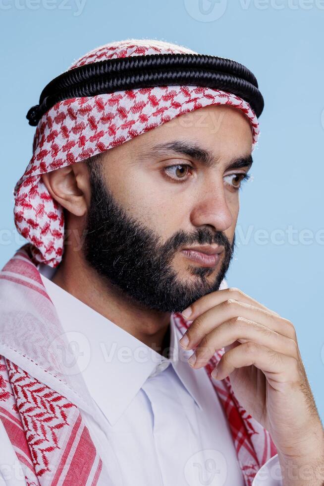 musulmano uomo nel tradizionale copricapo pensiero e considerando soluzione. arabo persona indossare ghutra foulard sfregamento mento e guardare lontano con concentrato espressione avvicinamento foto