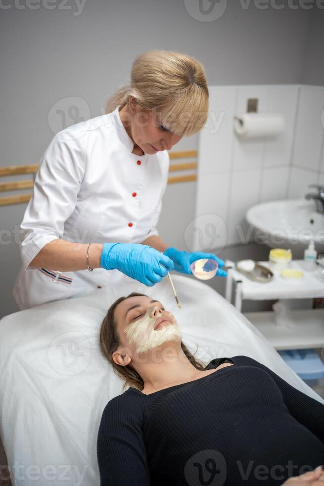 medico cosmetologo o dermatologo fabbricazione viso maschera nel cosmetologia salone. professionale estetista l'applicazione viso maschera su caucasico donna viso dire bugie su letto nel accappatoio. foto