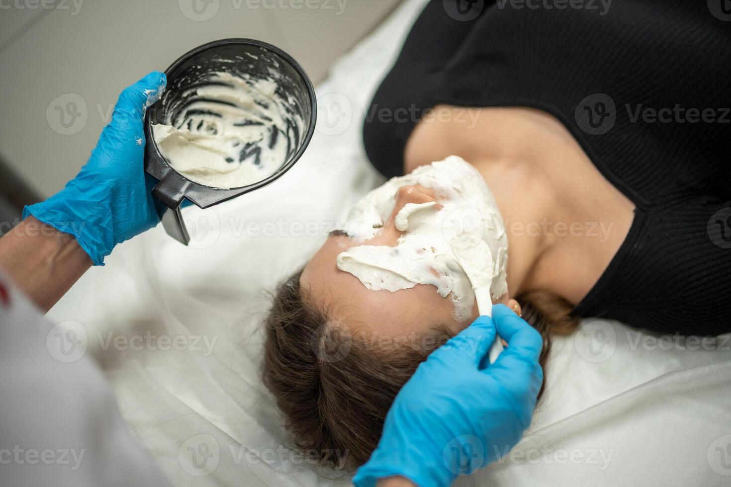 medico cosmetologo o dermatologo fabbricazione viso maschera nel cosmetologia salone. professionale estetista l'applicazione viso maschera su caucasico donna viso dire bugie su letto nel accappatoio. foto