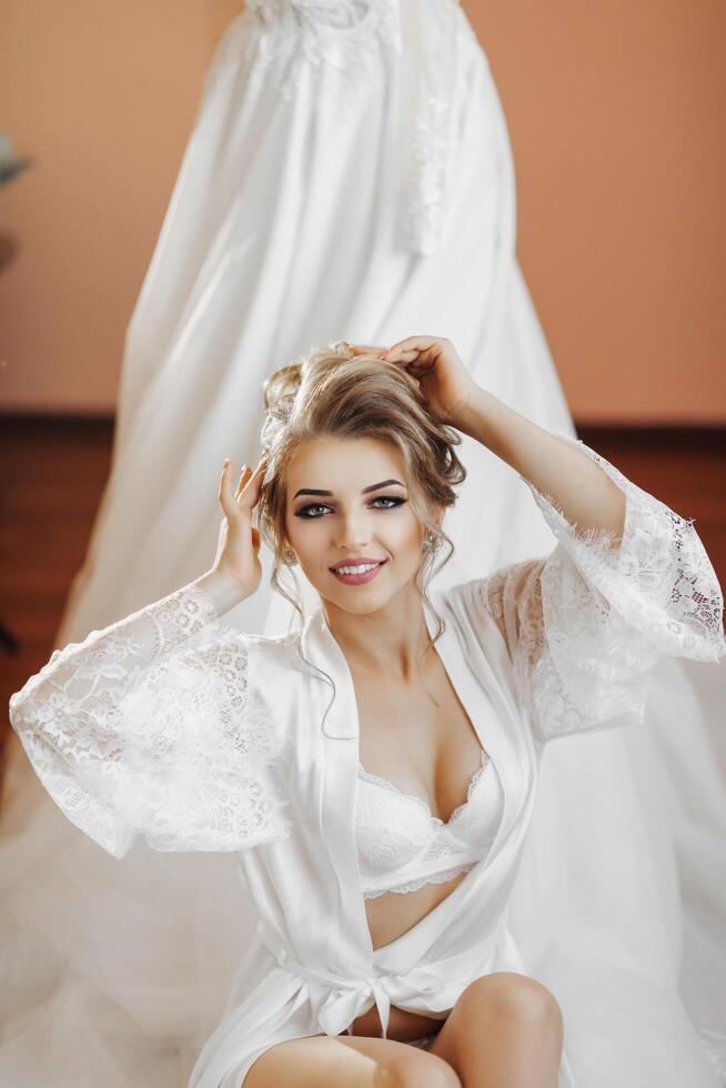 un' bionda sposa nel un' lungo pizzo accappatoio e bianca scarpe si siede nel sua camera Il prossimo per sua nozze vestire, in posa con sua braccia attraversato di sua testa. bellissimo capelli e trucco, Aperto rompersi. nozze ritratto. foto