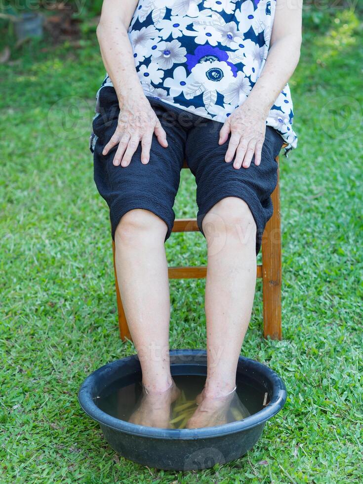 un' anziano donna ha un caviglia dolore trattamento di terme piede con erbe aromatiche nel il giardino. il muscoli di ammollo caldo acqua quello è bollito a partire dal erbe aromatiche. concetto di anziano persone e assistenza sanitaria foto