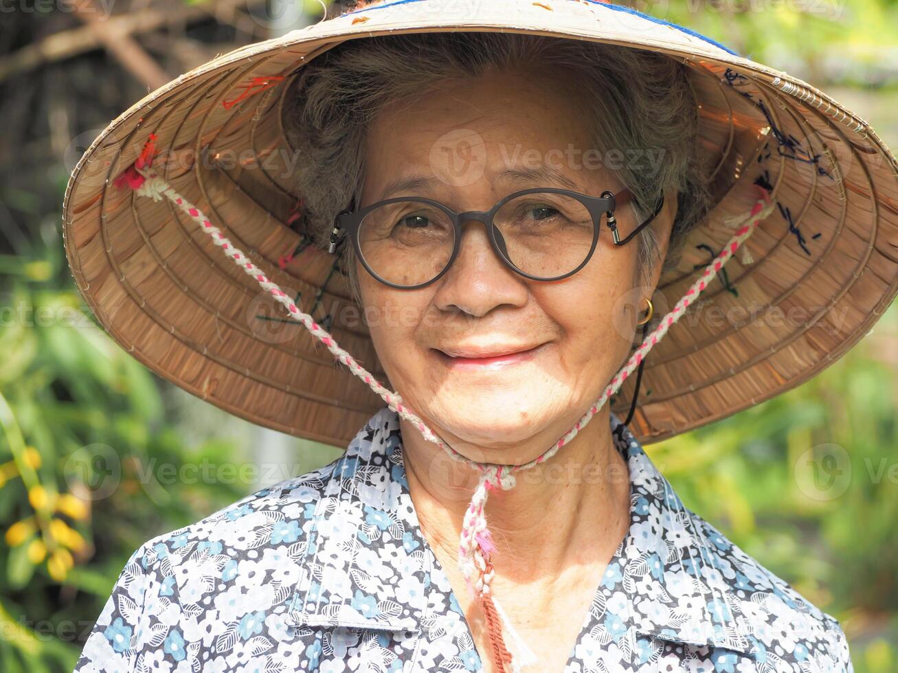 ritratto di un' anziano asiatico donna indossare un' cappello, sorridente e guardare a il telecamera mentre in piedi nel un' giardino. spazio per testo. concetto di vecchio persone e assistenza sanitaria foto