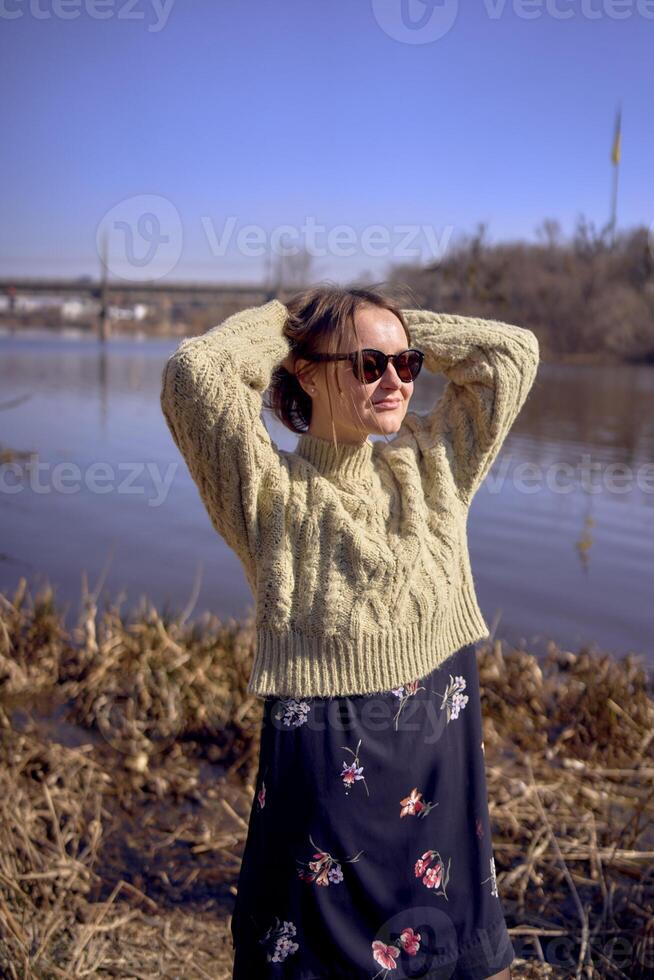 elegante giovane donna nel un' leggero verde maglione e occhiali da sole su il banca di un' primavera fiume foto