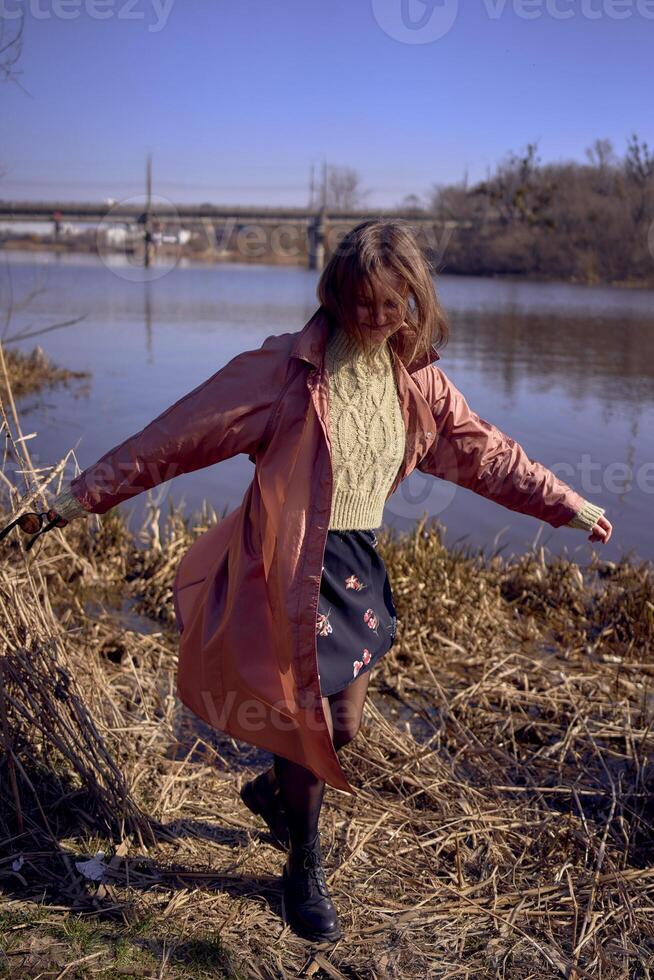 un' elegante giovane donna nel un' rosa trincea balli su il banca di un' primavera fiume foto
