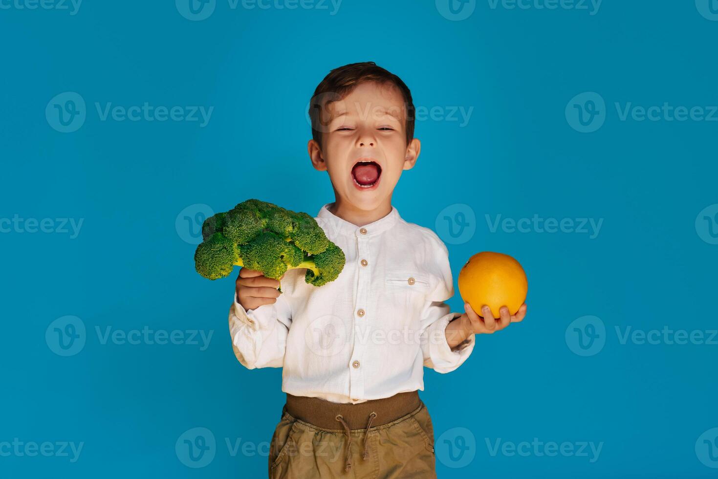 un' studio tiro di un' ragazzo Tenere fresco broccoli e un arancia su un' blu sfondo. il concetto di salutare bambino cibo. foto