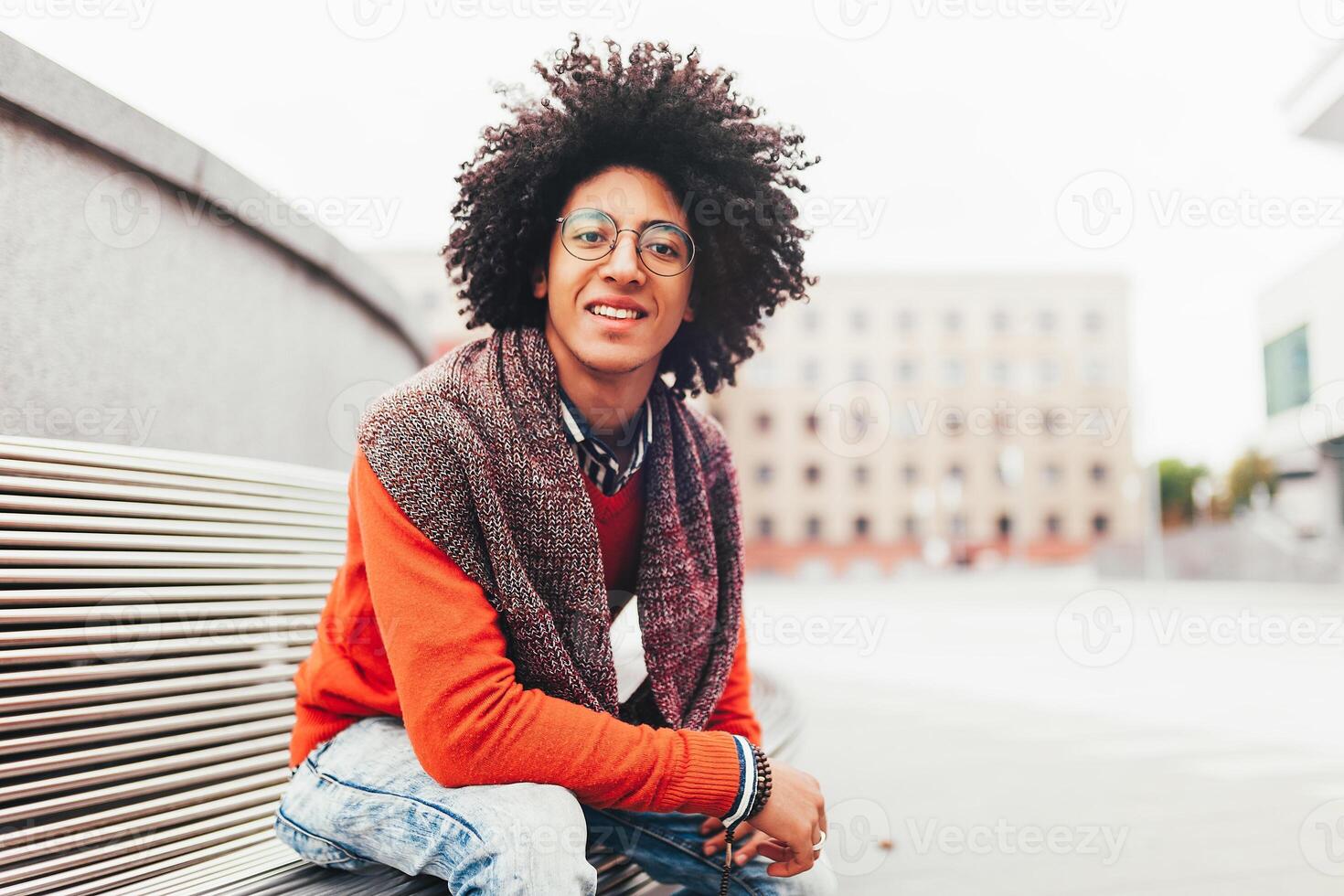 un' bello giovane egiziano Riccio tipo seduta su un' panchina vestito nel un' luminosa arancia maglione e jeans. contento le persone. il alunno passeggiate nel il rompere fra il Lezioni. foto