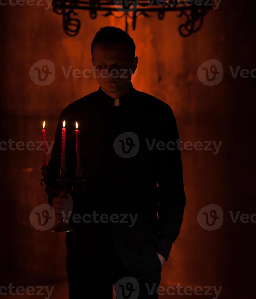 giovane cattolico preghiere sacerdote. ritratto di sacerdote Il prossimo per il candele prega con il suo mani piegato vicino il viso. rosso parete su il sfondo. interno bello ritratto di religione uomo foto