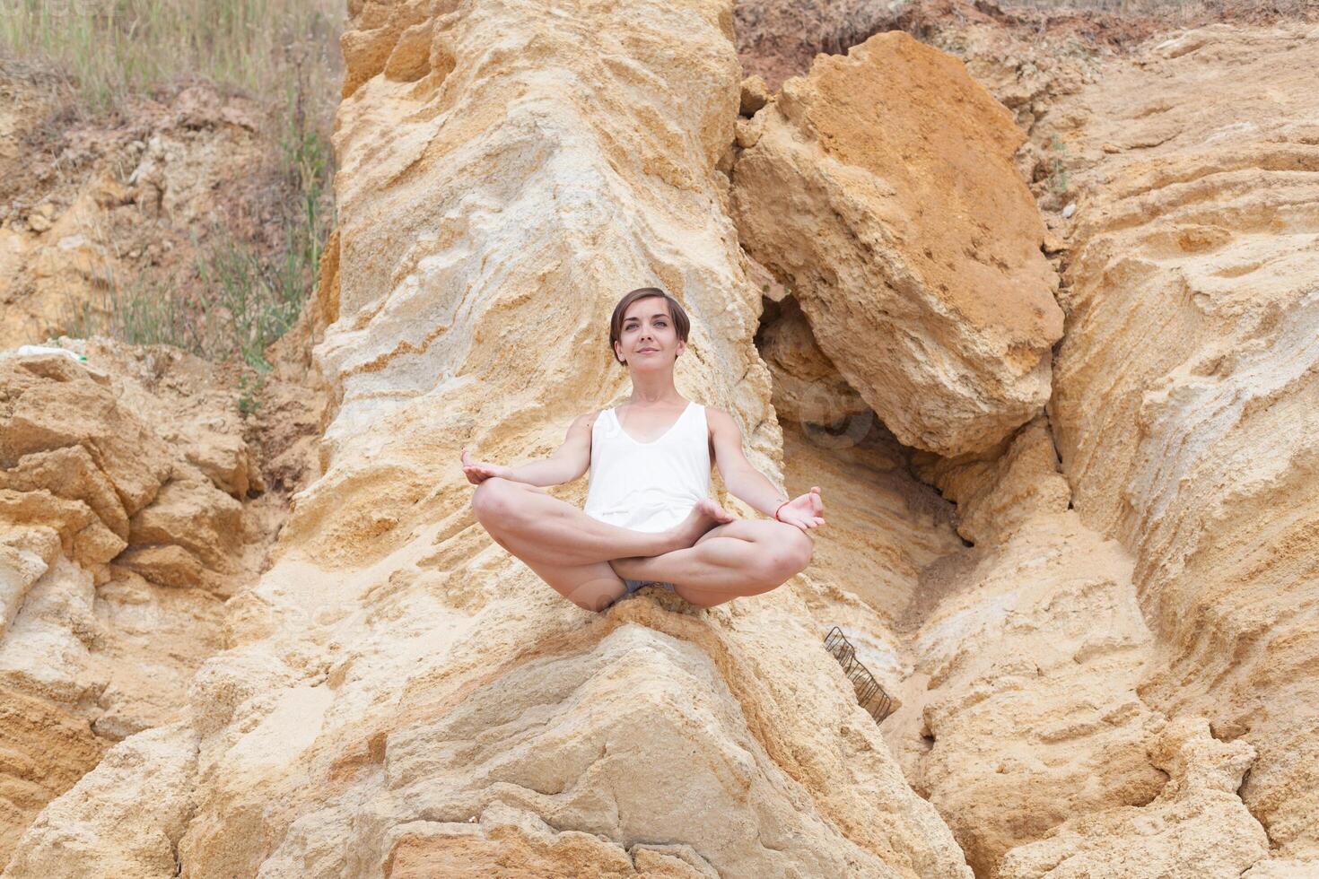 un' bellissimo giovane ragazza con corto capelli è vestito nel pantaloncini e un' bianca maglia è praticante yoga su il sfondo di rocce. posa di il loto. il concetto di calma e concentrazione foto