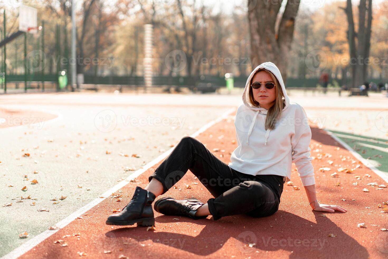 all'aperto ritratto di giovane bellissimo donna con lungo nel occhiali da sole e un' bianca incappucciato maglione seduta su il campo sportivo traccia foto
